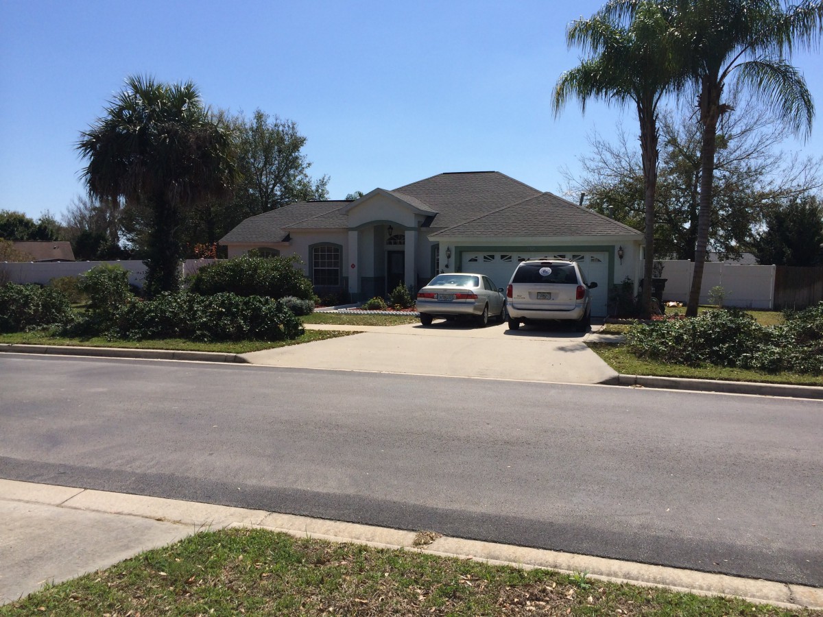 A suburban house with a driveway and two cars parked in front, surrounded by palm trees on a sunny day.