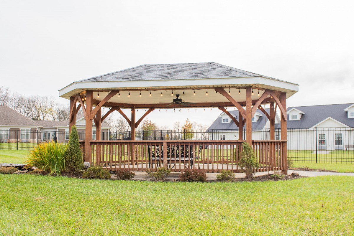 A wooden gazebo stands in a garden surrounded by green lawn and houses.