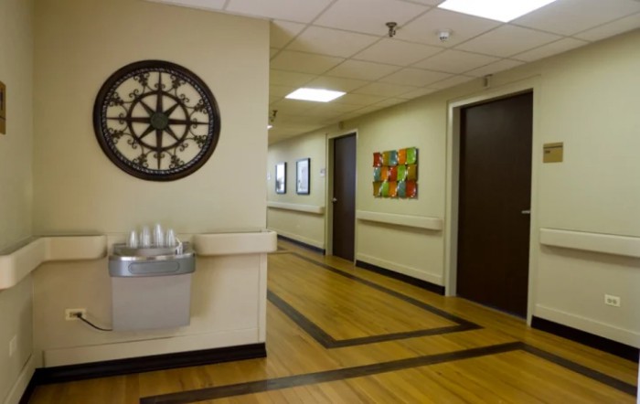 A hallway with wooden flooring, featuring a decorative wall clock, artwork on the walls, and a water fountain.