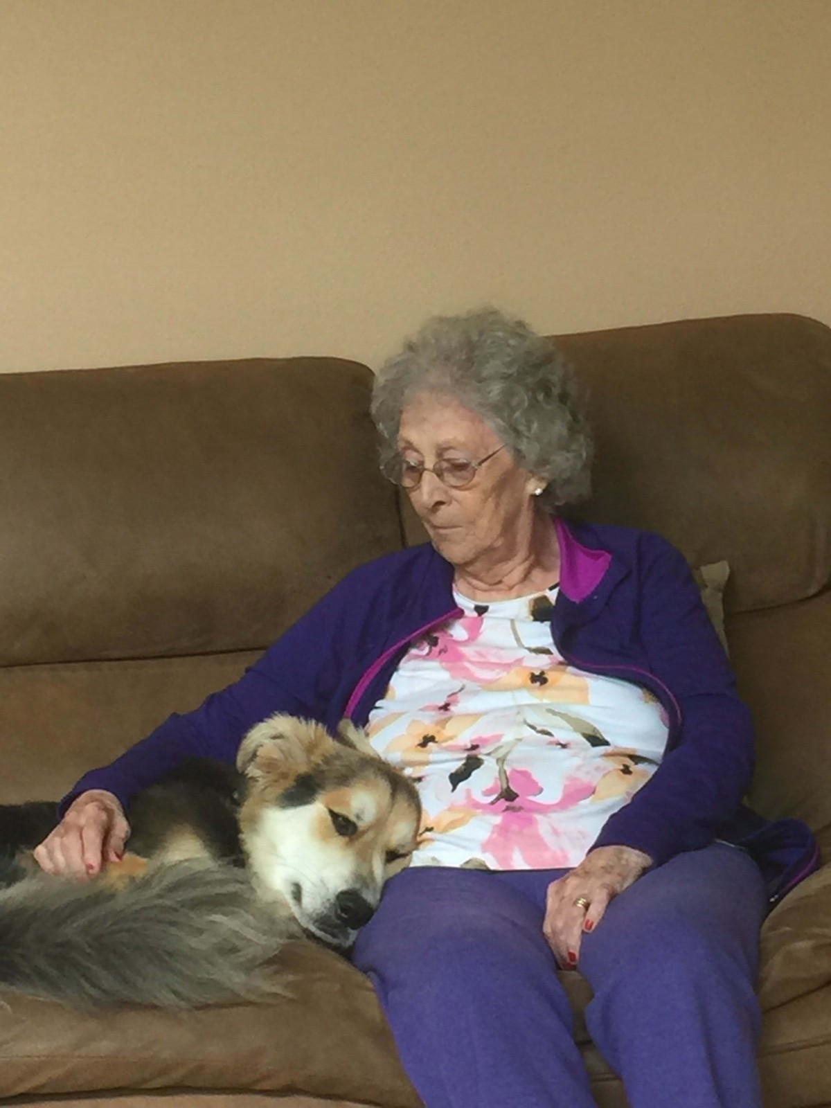 An elderly woman sitting on a couch with a dog resting beside her.