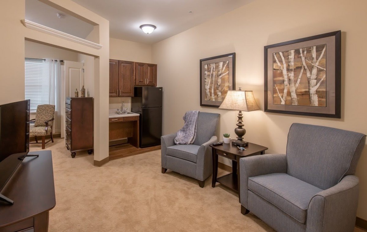 A cozy living room with two chairs, a lamp, and wall art near a small kitchenette.