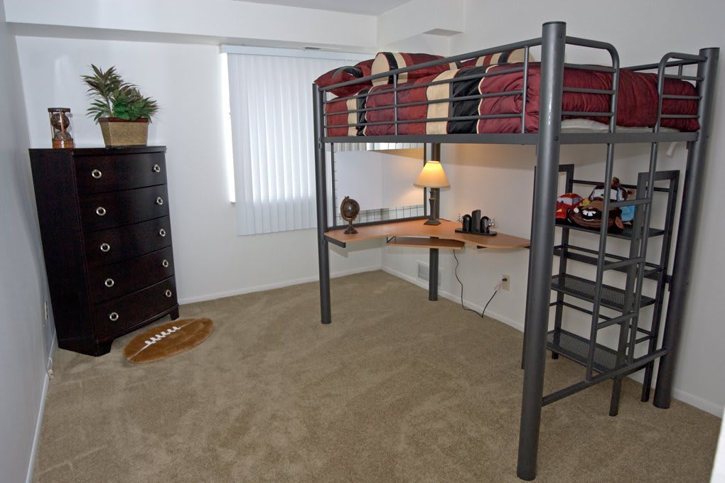 A minimalist bedroom featuring a loft bed with an attached desk, a lamp, and a dresser.