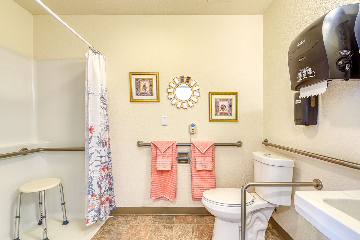 A clean and well-decorated bathroom featuring a toilet, shower area with a curtain, wall decorations, and pink towels hung on grab bars.