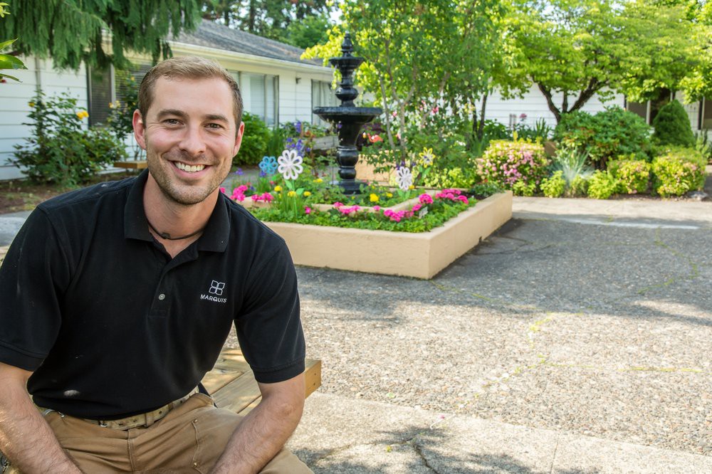 A man smiling in a well-maintained garden with a fountain and blooming flowers.