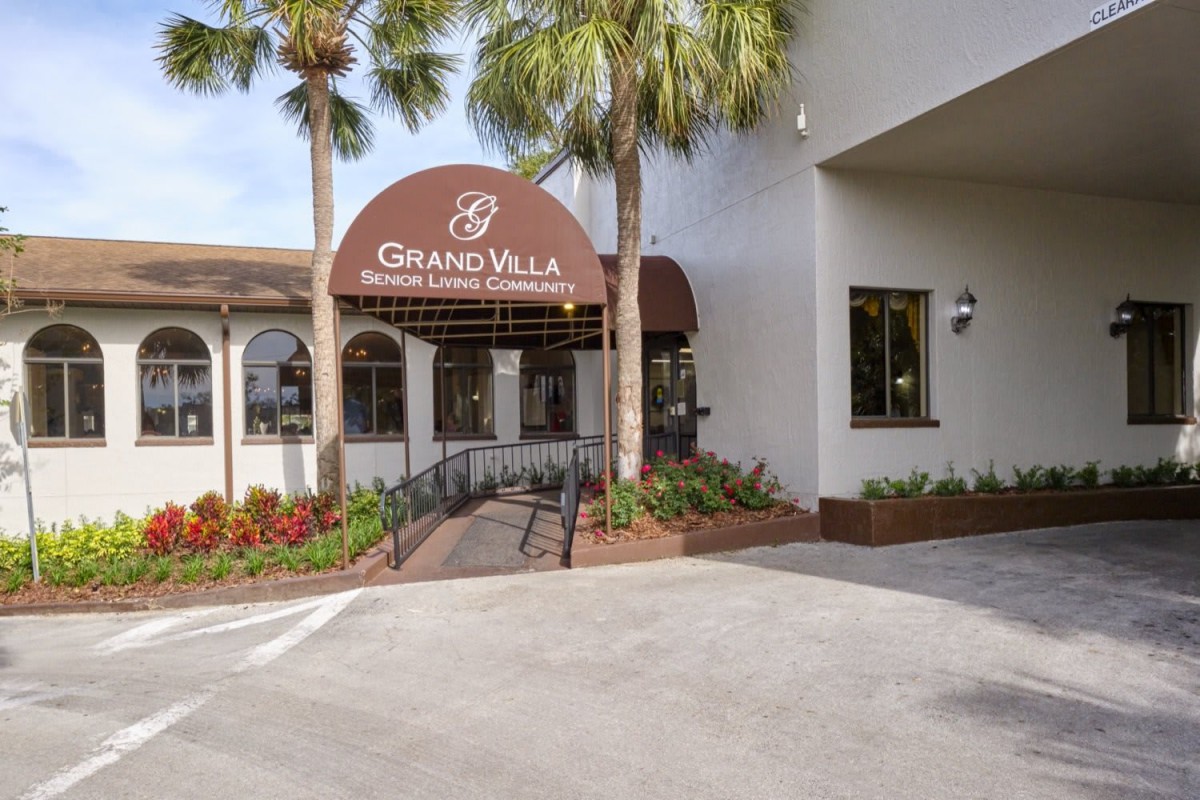 Entrance to the Grand Villa Senior Living Community with palm trees and well-maintained landscaping.
