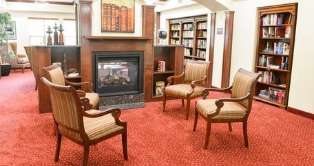 A cozy library setting with armchairs around a fireplace and bookshelves in the background.