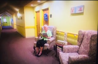 An elderly woman sits in a hallway reading a magazine, surrounded by homey decor.