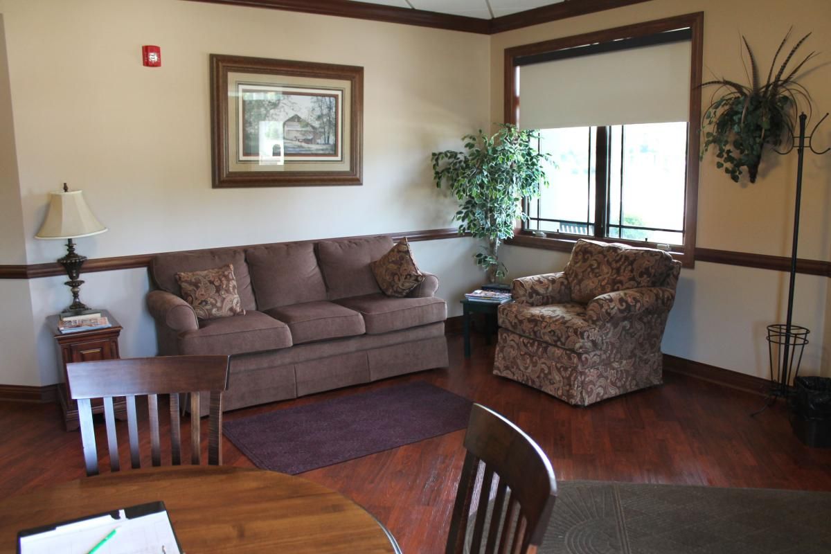 A cozy living room with a brown sofa, patterned armchair, and a coffee table, featuring an indoor plant and large windows.