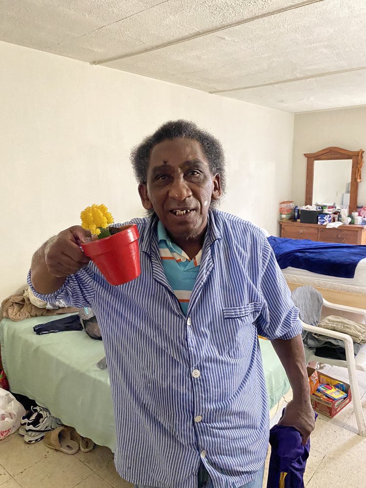 An elderly woman holding a red pot with a yellow flower inside, standing in her living space.
