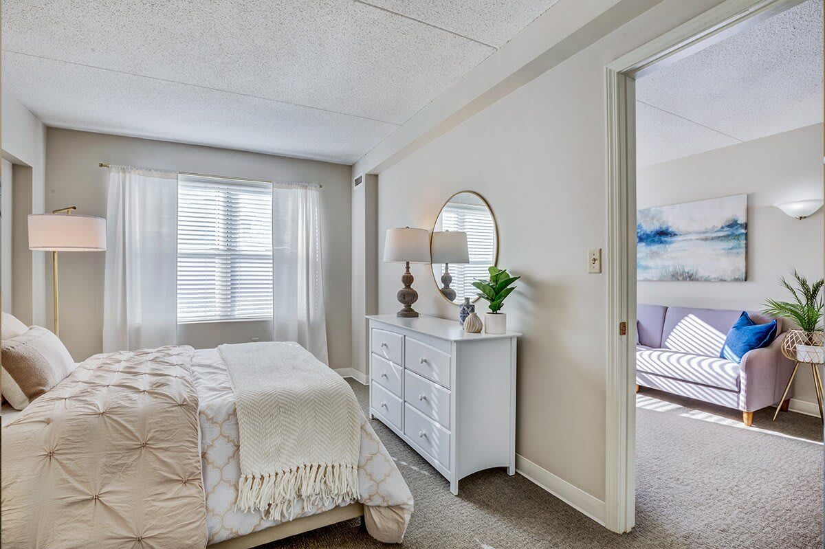 A serene bedroom featuring a cozy bed, a white dresser with a round mirror, and soft natural light filtering through sheer curtains.