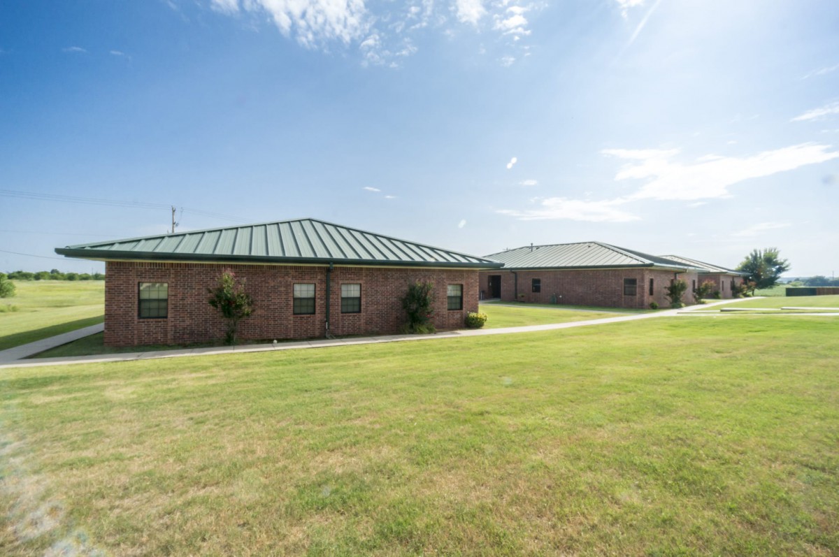 Two brick buildings with green roofs surrounded by a grassy lawn under a blue sky.
