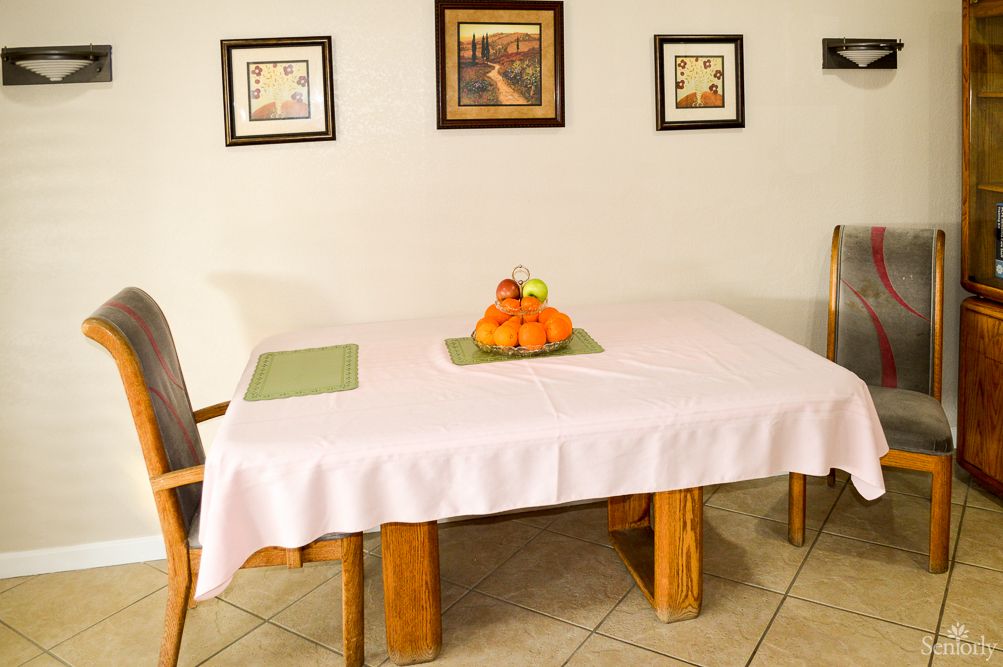 A dining room with a white tablecloth-covered table, adorned with a fruit basket and surrounded by chairs. The walls are decorated with framed artwork.