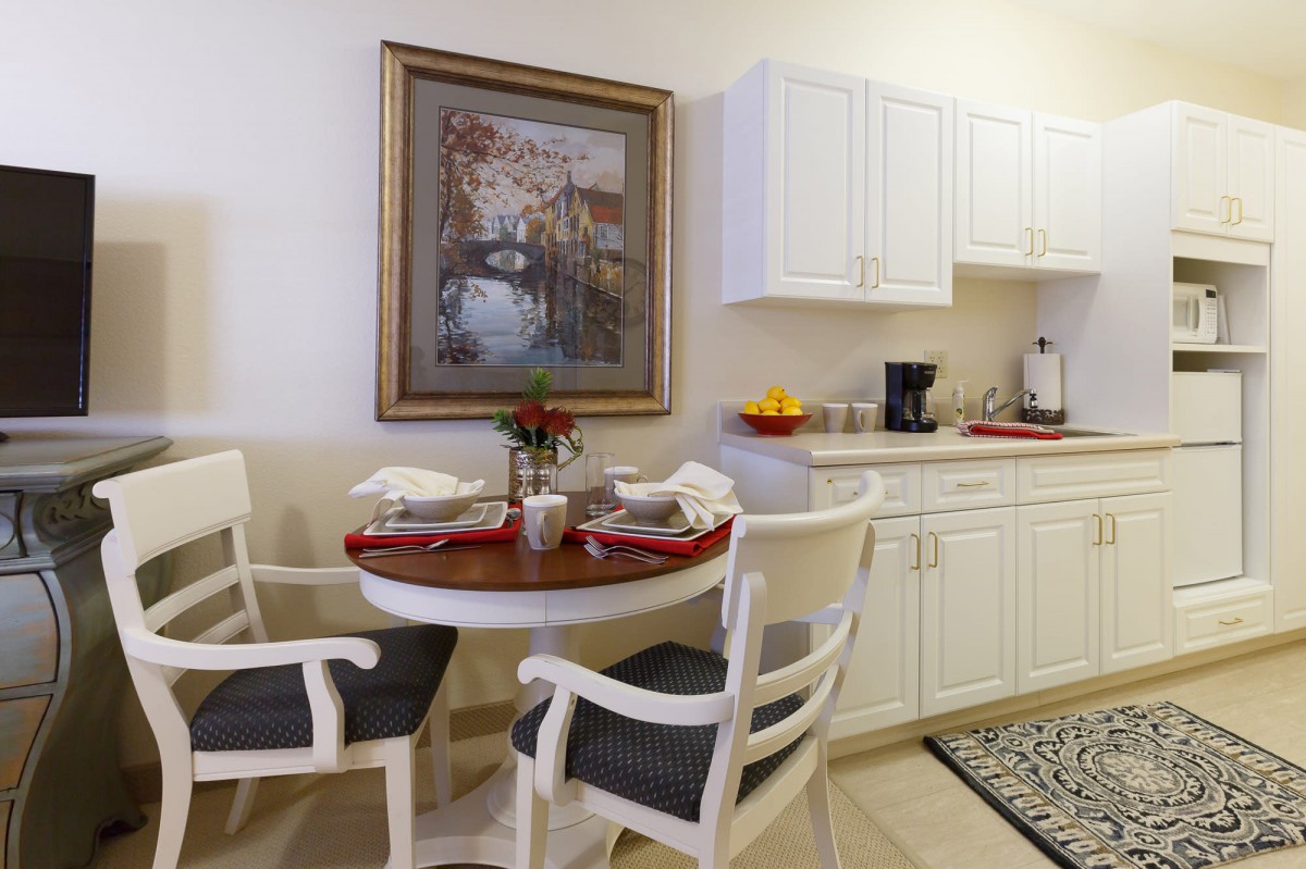 A cozy kitchen and dining area with white cabinetry, a round table set for two, and a decorative painting on the wall.
