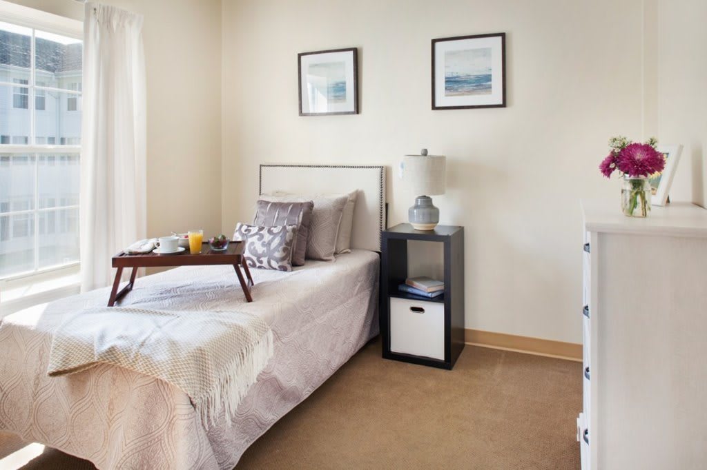 A modern bedroom with a neatly made bed, decorative pillows, and a tray on top. The room features a nightstand with a lamp and framed artwork on the walls.