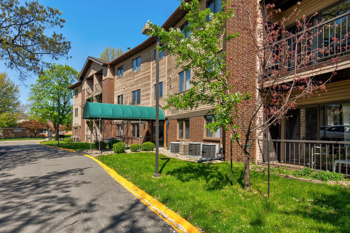 A three-story apartment building with a brick exterior, surrounded by greenery on a sunny day.
