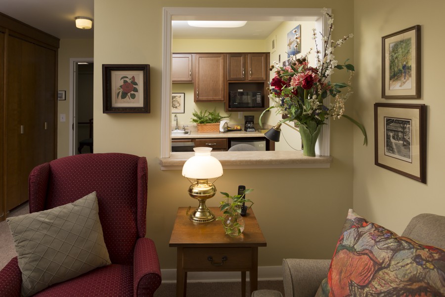 A cozy living room with a view into the kitchen, featuring comfortable furniture, a lamp, and a floral arrangement.