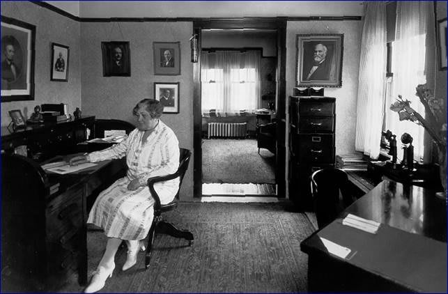 A vintage black and white photo of a woman sitting at a desk in an office, surrounded by portraits on the walls.