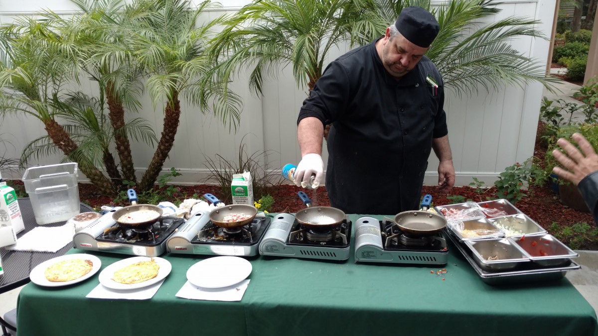 A chef preparing food outdoors using portable stoves surrounded by palm trees.