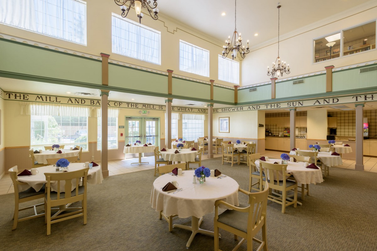 A spacious and elegantly designed dining room with round tables, wooden chairs, and large windows allowing natural light to fill the space.