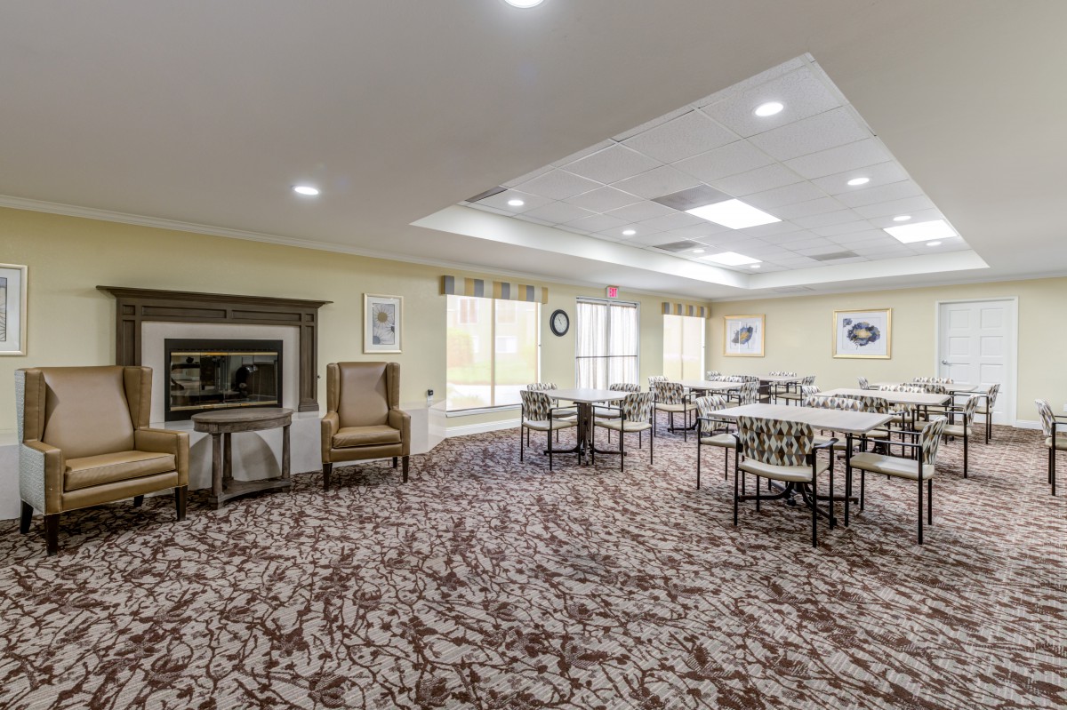 A spacious dining area with several tables and chairs, featuring a patterned carpet, large windows, and a fireplace.