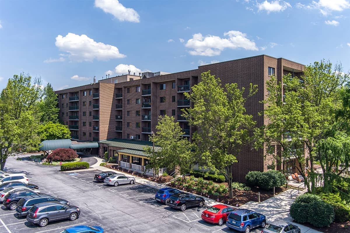 A multi-story apartment complex with a parking lot full of cars, surrounded by trees under a partly cloudy sky.