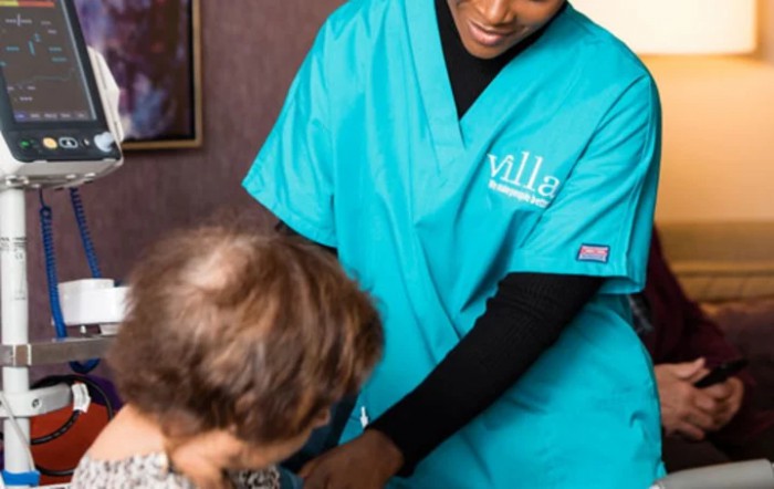 A nurse in teal scrubs assists an elderly person.