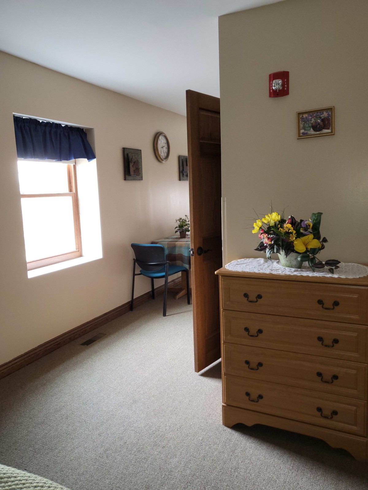 A cozy room with a dresser topped with flowers, a window, and a chair in the corner.