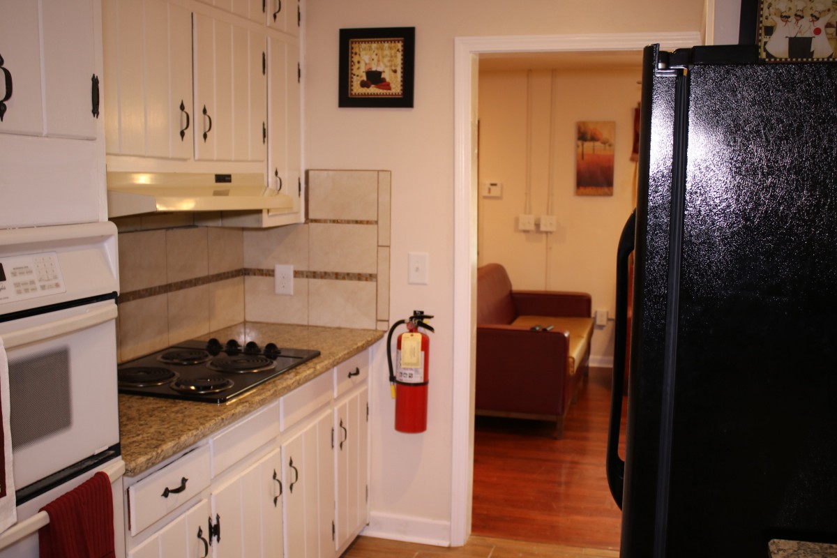A view of a kitchen with cream cabinets and a black refrigerator, leading to a living room with a brown couch.