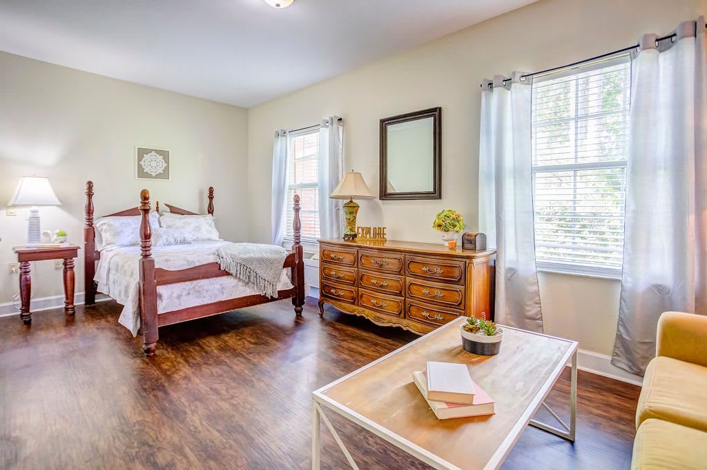 A bright bedroom with wooden flooring, featuring a bed, dresser, and ample natural light.