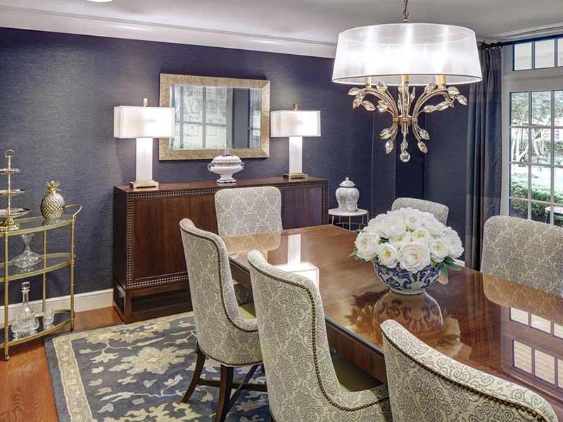 An elegantly designed dining room featuring a large wooden table, upholstered chairs, and a chandelier. The walls are adorned with a mirror and decorative lighting.