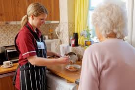 A caregiver assists an elderly person by preparing a meal in the kitchen.