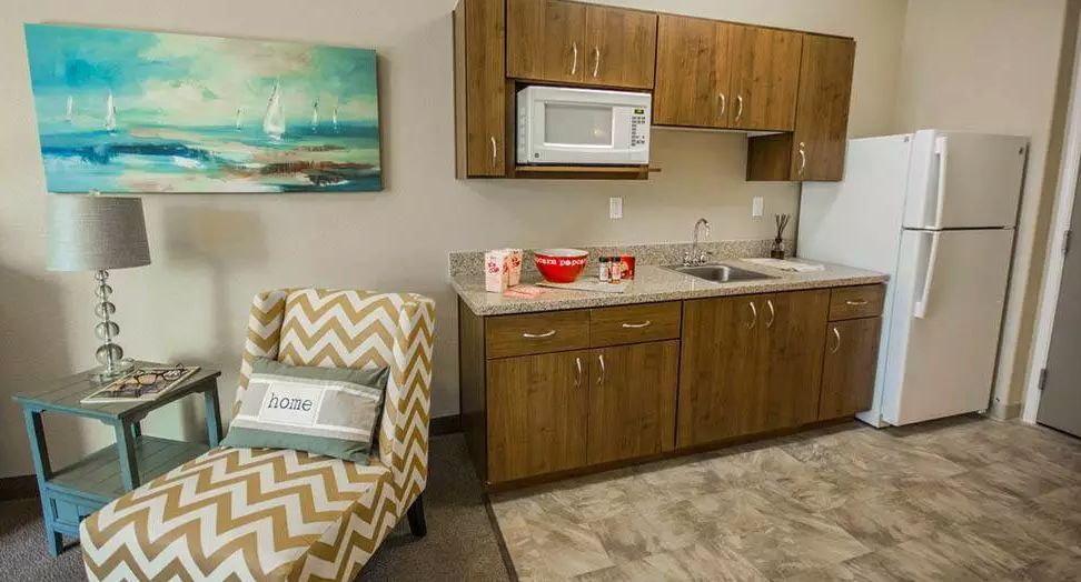 A compact kitchenette area with wooden cabinets, a microwave, and a sink, adjacent to a patterned armchair.