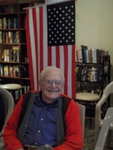 A person sitting indoors with an American flag and a bookshelf in the background.