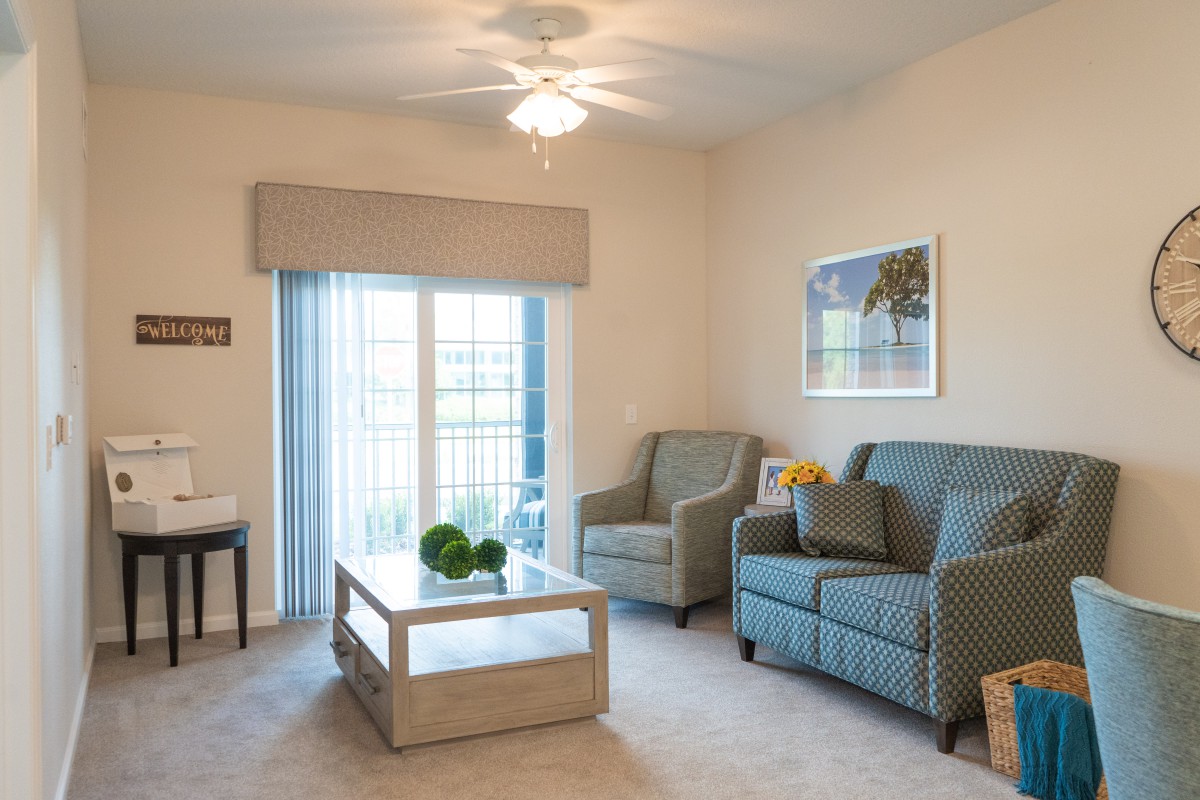 A cozy living room with a sofa and armchair, featuring a central coffee table and a large window with curtains.