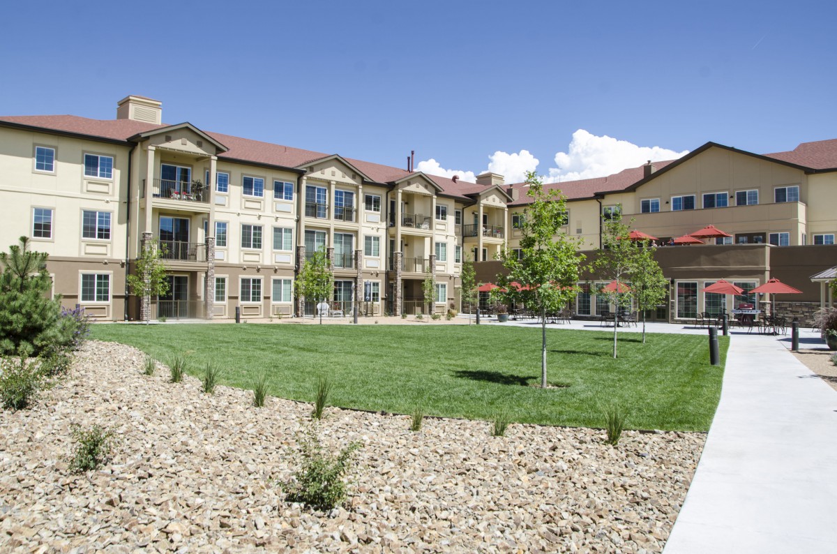 A three-story apartment building with a well-maintained lawn and landscaped surroundings under a clear blue sky.