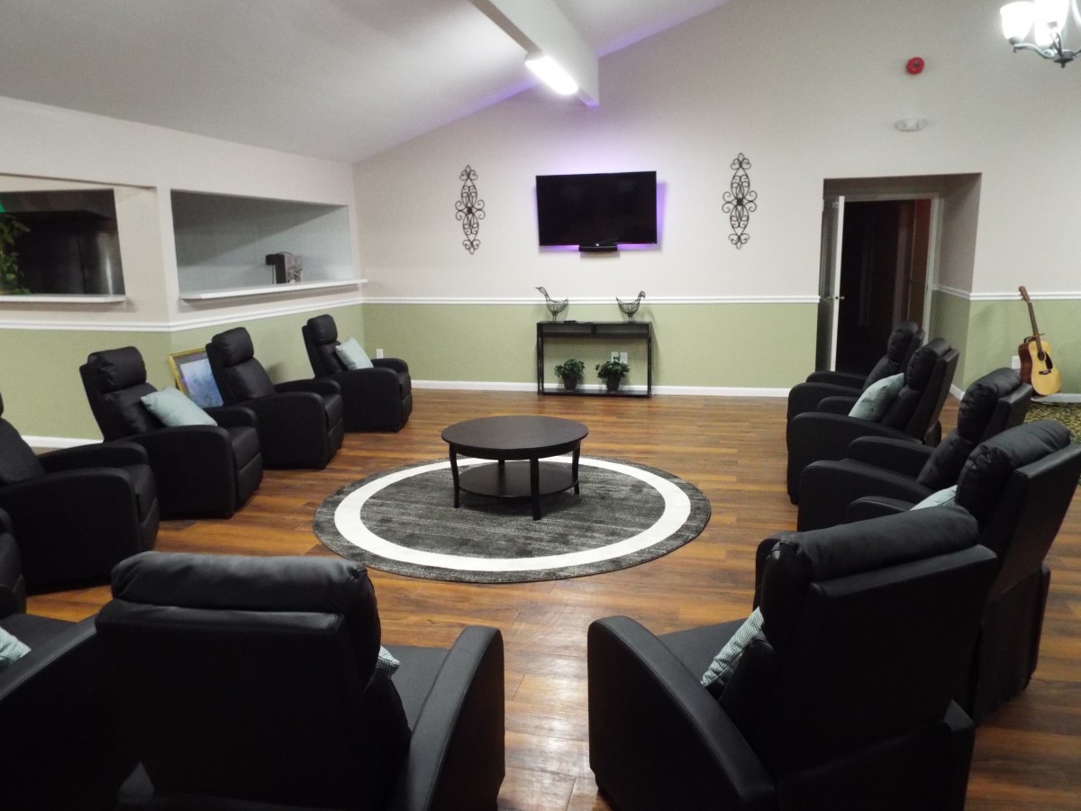 A cozy seating area with armchairs arranged in a circle around a central table, featuring a television on the wall and wooden flooring.