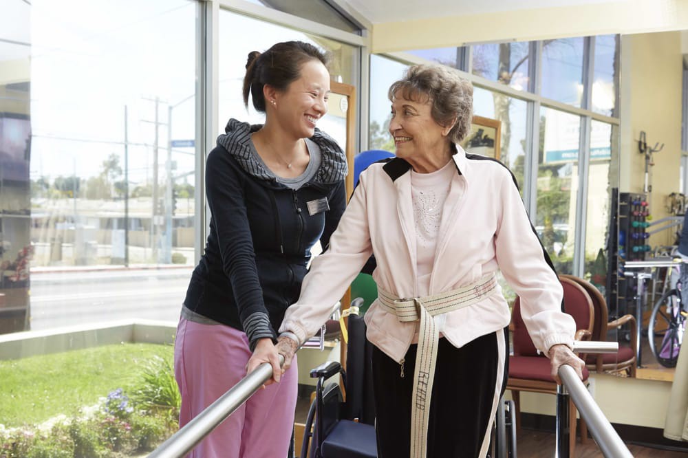 A caregiver assists an elderly woman during a physical therapy session.