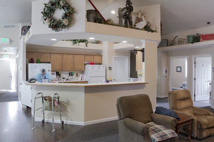 A cozy living area with a view of the kitchen, featuring a wreath decoration on the wall, comfortable seating, and mobility aid equipment.