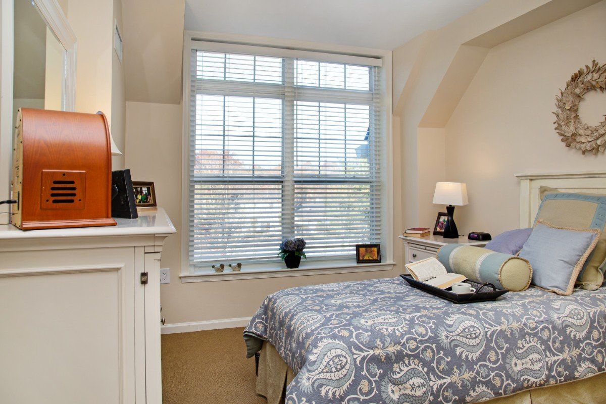 A cozy bedroom with a large window, featuring a bed with patterned bedding and a small lamp on the nightstand.