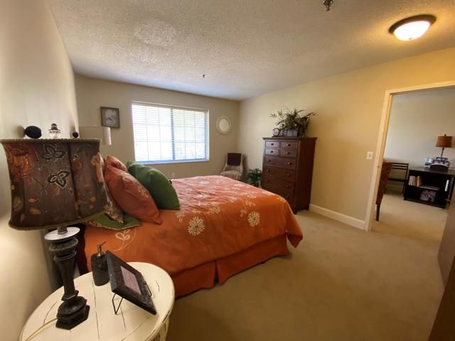 A cozy bedroom with warm tones, featuring a bed with orange bedding and green pillows, a lamp on the side table, and a window letting in natural light.