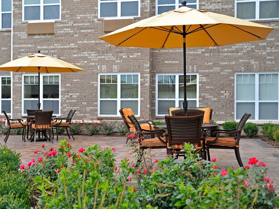A serene outdoor patio area with tables, chairs, and umbrellas in front of a brick building, surrounded by flowers.