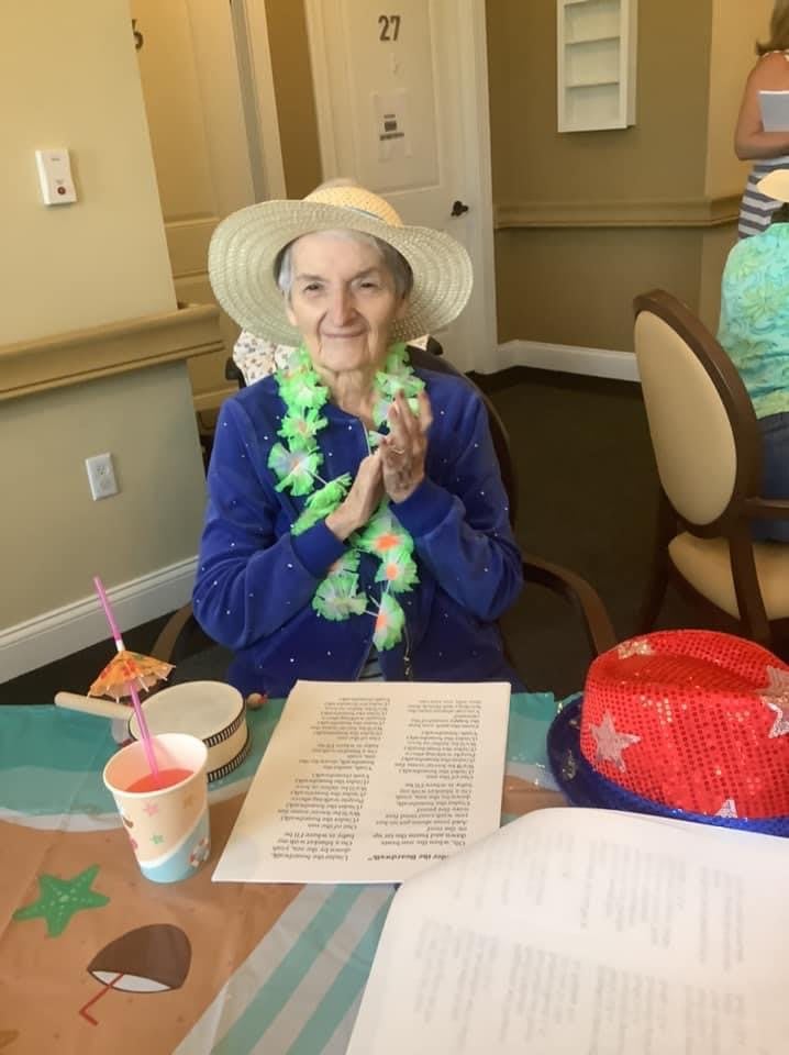 An elderly woman wearing a hat and leis sits at a table during a celebration.