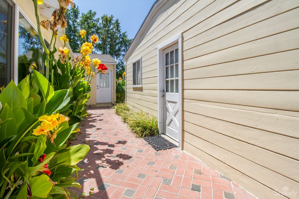 A sunny walkway beside a house, lined with colorful flowers and greenery.