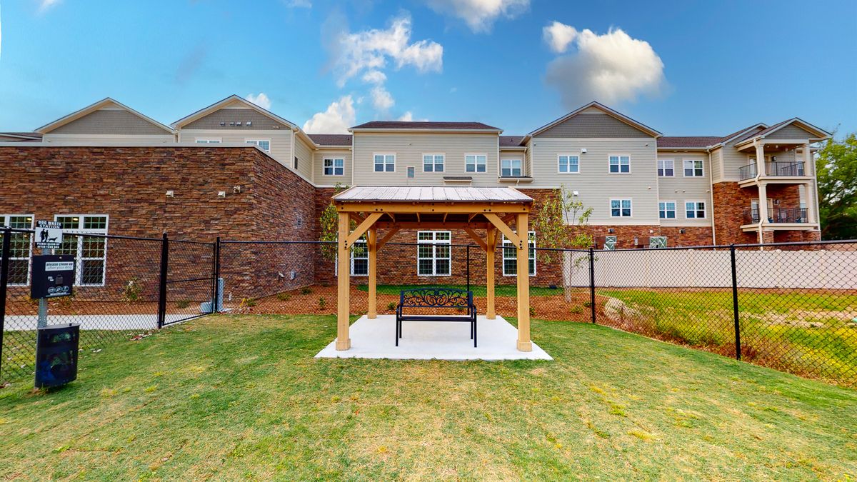 A small wooden gazebo with a swing sits on a grassy lawn in front of a large multi-story building under a partly cloudy sky.