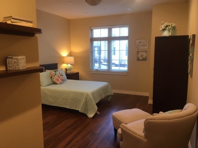 A cozy bedroom with soft lighting, featuring a bed, chair, and a window for natural light.
