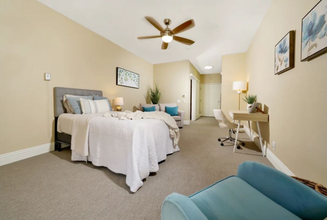 A spacious bedroom featuring a double bed, modern decor, and a ceiling fan. The room includes a desk with a lamp and is decorated in neutral tones.