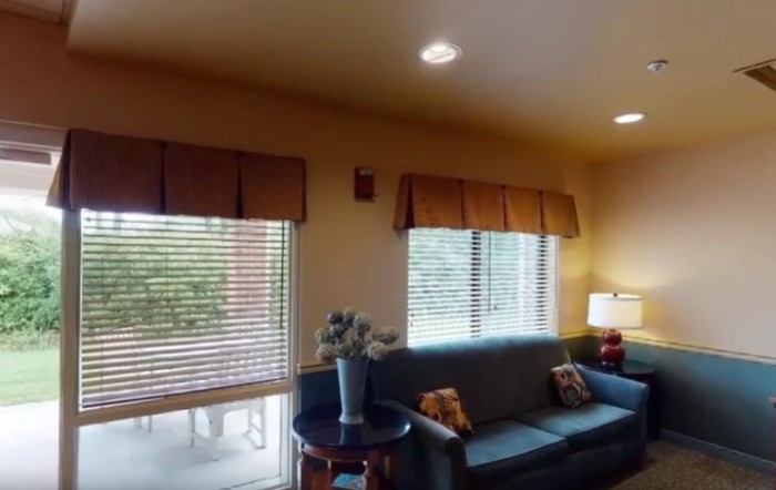 A cozy living room with a sofa, windows with blinds, and a side table holding a vase of flowers.
