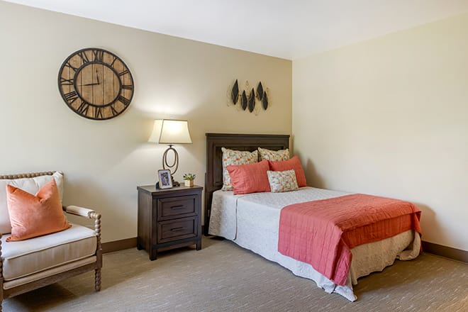 A cozy bedroom with warm decor, featuring a bed with coral accents, a wooden nightstand with a lamp, and a decorative wall clock.