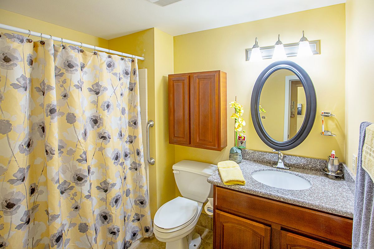 A bright bathroom with yellow walls, featuring a floral shower curtain, oval mirror, wooden cabinet, and standard fixtures.