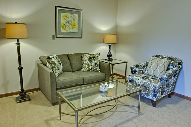A cozy living room setup with a grey sofa, patterned armchair, and glass coffee table next to a stylish lamp and framed artwork.